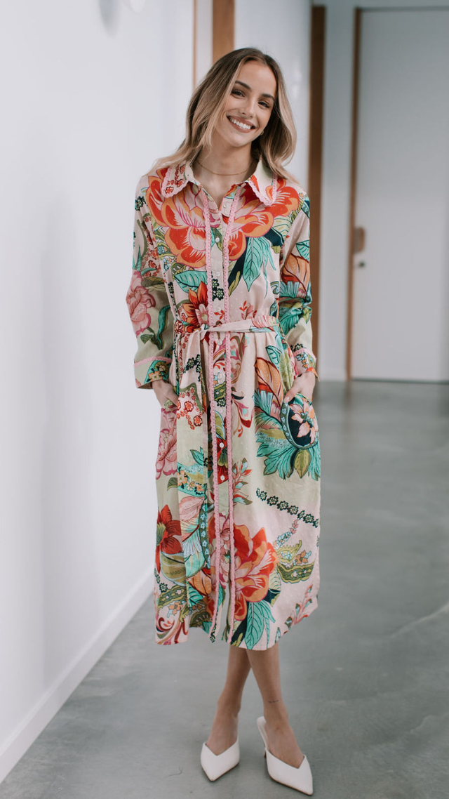 Woman smiling in the Medusa Dress, a floral button-down shirt dress with waist tie, long sleeves, and a vibrant tropical print.