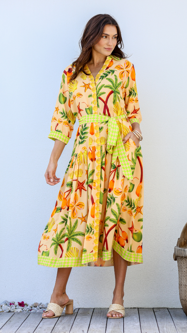 Model wearing the Diego Dress – a vibrant button-down midi dress with a tropical fruit and shell print, green check belt, and neon trim detailing.