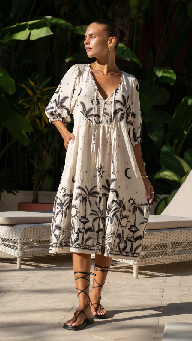 Model wearing the Hattie Dress in Cosmic – midi length ivory dress with black celestial palm and moon print, tie-front neckline, puff sleeves, side pockets, and relaxed flowy silhouette.