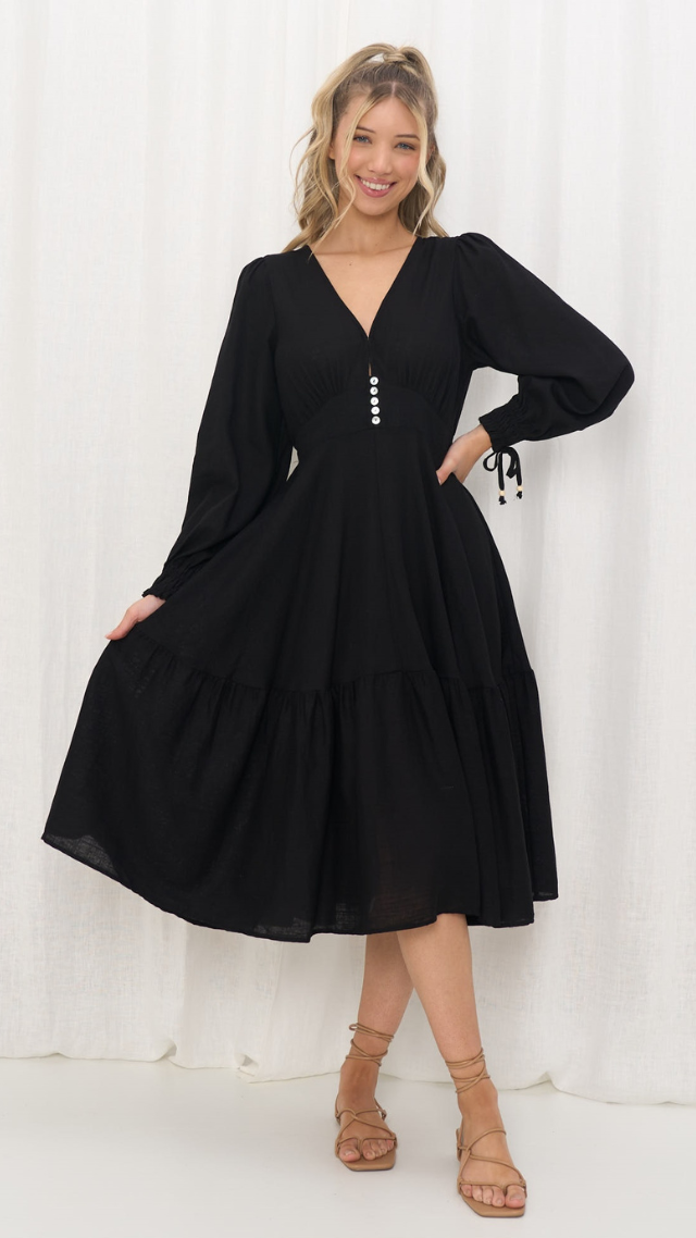 Model wearing the Margo Dress in black, featuring a V-neckline with buttons, long balloon sleeves with tie cuffs, and a tiered A-line midi skirt.