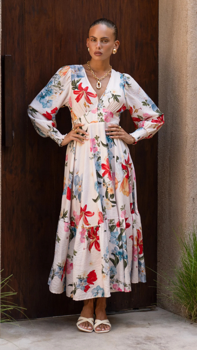 Model wearing the Parsons Long Sleeve Dress in Lily print – a V-neck, long sleeve midi dress with tiered skirt, smocked cuffs, and vibrant floral pattern on ivory base.
