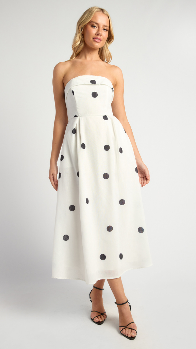 Model wearing the Spot On Dress in white with black polka dots – strapless, fitted bodice with full pleated midi skirt and bold retro-inspired print.
